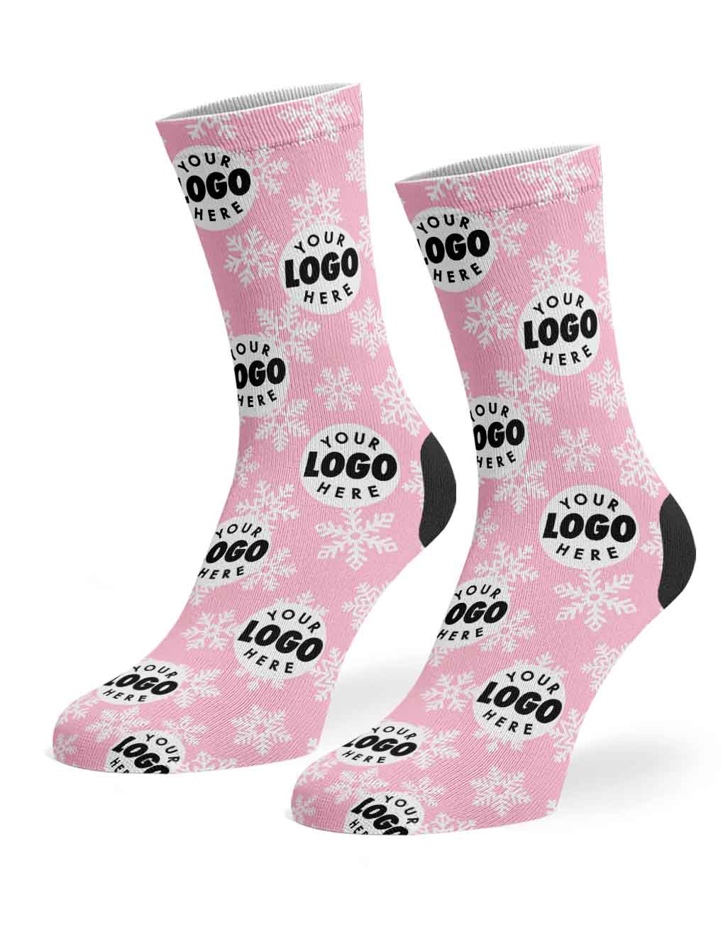 A pair of pink crew socks with a white holiday knit pattern featuring snowflakes, pine trees, and black placeholder text reading 'YOUR LOGO HERE', shown on a white background.